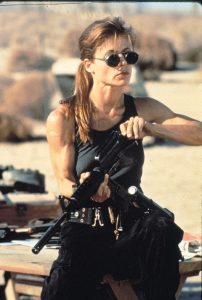 Linda Hamilton in scene from Terminator 2 sitting on table loading machine gun wearing sunglasses